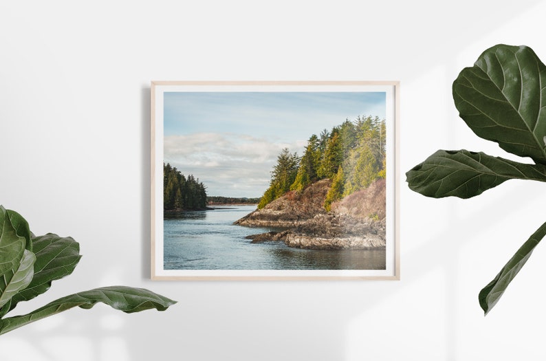 May include: A framed landscape print featuring a coastal scene. The image shows a body of water surrounded by rocky cliffs and lush green trees under a cloudy sky. The frame is a light wood color.