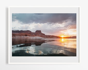 May include: A framed landscape photograph shows a sunset over a lake. The sky is filled with dramatic clouds, reflected in the calm water. The sun's golden light illuminates the scene, highlighting the rocky formations in the distance.