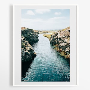May include: A narrow, winding river flows through a rocky canyon. The water is a deep blue, and the rocks are a mix of brown, green, and gray. The sky is a light blue with a few white clouds.