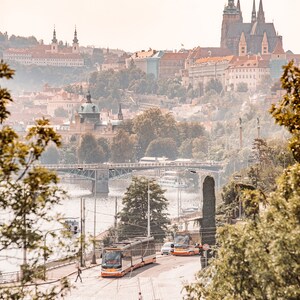 May include: A view of Prague, Czech Republic, with a cityscape in the background and two orange trams in the foreground. The trams are on a street with a river and a bridge in the distance.