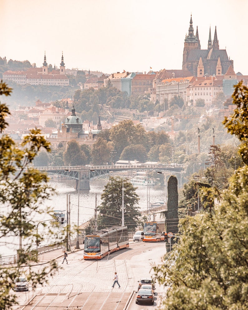 May include: A view of Prague, Czech Republic, with a cityscape in the background and two orange trams in the foreground. The trams are on a street with a river and a bridge in the distance.