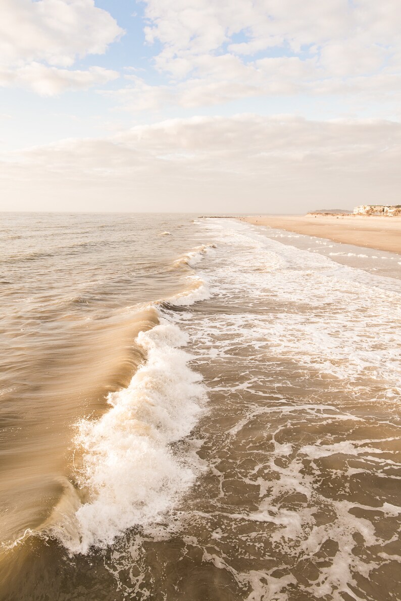 Pu&ograve; includere: Una serena scena di spiaggia con un'onda che si infrange in primo piano. L'acqua &egrave; di un marrone tenue, in contrasto con la schiuma bianca. Il cielo &egrave; azzurro pallido con nuvole soffici e la sabbia &egrave; beige chiaro.