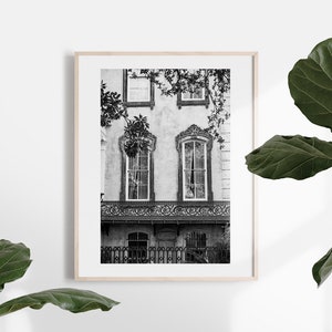 May include: Black and white framed print of a historic building facade. The image features ornate windows, a decorative balcony, and a wrought-iron fence. The building is partially obscured by tree branches, adding depth to the composition. The print is displayed in a light wood frame.