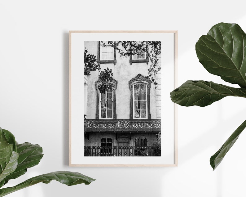 May include: Black and white framed print of a historic building facade. The image features ornate windows, a decorative balcony, and a wrought-iron fence. The building is partially obscured by tree branches, adding depth to the composition. The print is displayed in a light wood frame.