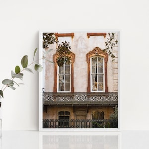 May include: A framed print of a historic building facade. The building features two arched windows with ornate trim, a wrought-iron balcony, and a weathered exterior. The color palette includes beige, brown, and white.