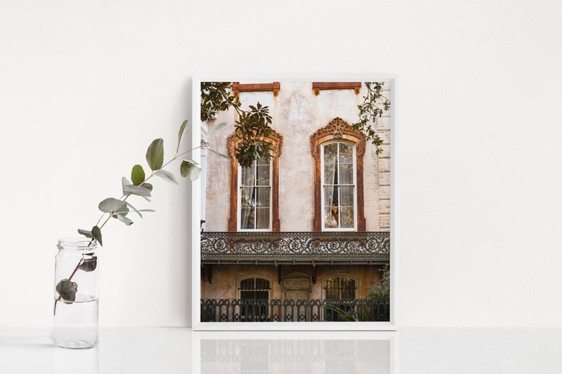 May include: A framed print of a historic building facade. The building features two arched windows with ornate trim, a wrought-iron balcony, and a weathered exterior. The color palette includes beige, brown, and white.