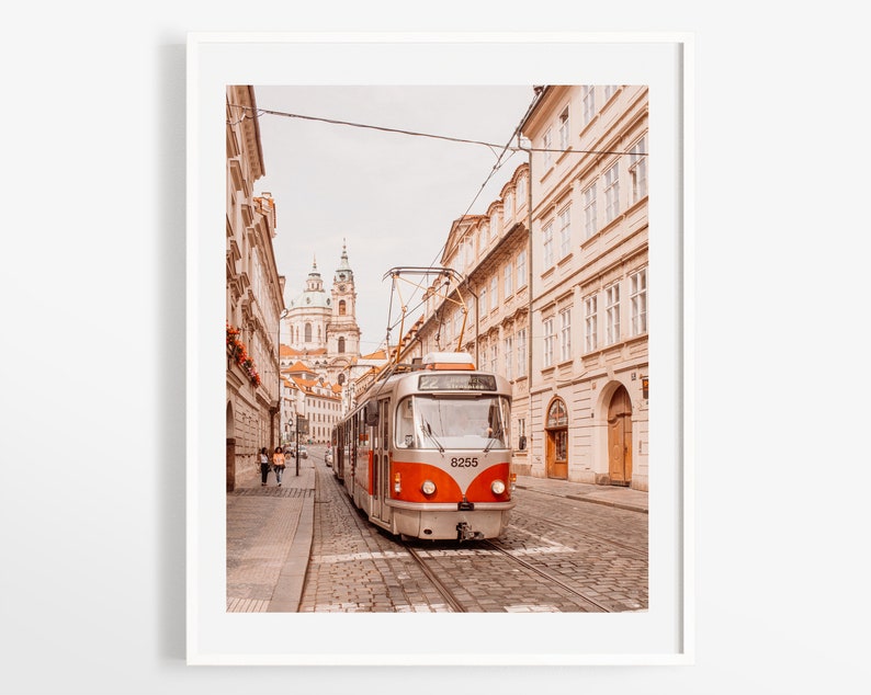 May include: A silver and orange tram travels down a cobblestone street in Prague, Czech Republic. The tram is numbered 8255. The street is lined with tall, pale yellow buildings. A church with a green dome is visible in the distance.
