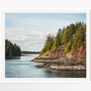 May include: A framed landscape print featuring a coastal scene. The image shows a body of water surrounded by rocky cliffs and lush green trees under a cloudy blue sky. The print is in a white frame.