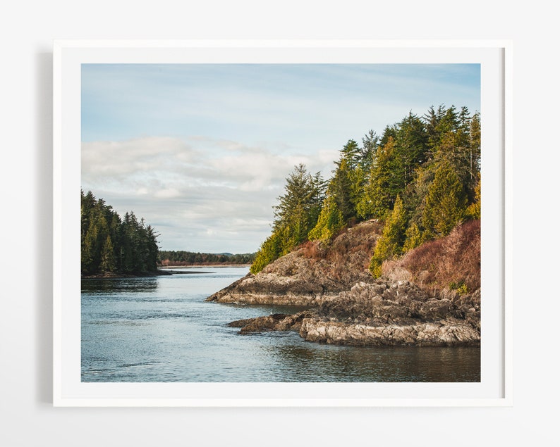 May include: A framed landscape print featuring a coastal scene. The image shows a body of water surrounded by rocky cliffs and lush green trees under a cloudy blue sky. The print is in a white frame.
