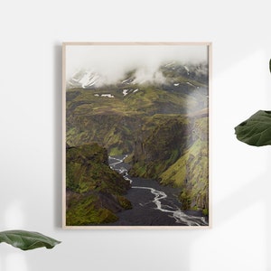 May include: A framed landscape print featuring a river winding through a deep canyon. The canyon walls are covered in green vegetation, with snow-capped mountains in the background. The print is in a light wood frame.