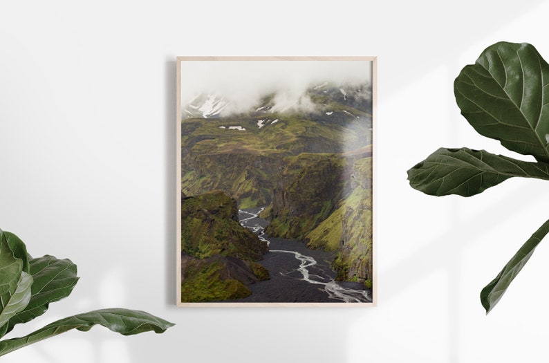 May include: A framed landscape print featuring a river winding through a deep canyon. The canyon walls are covered in green vegetation, with snow-capped mountains in the background. The print is in a light wood frame.