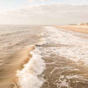 Pu&ograve; includere: Una serena scena di spiaggia con un'onda che si infrange in primo piano. L'acqua &egrave; di un marrone tenue, in contrasto con la schiuma bianca. Il cielo &egrave; azzurro pallido con nuvole soffici e la sabbia &egrave; beige chiaro.