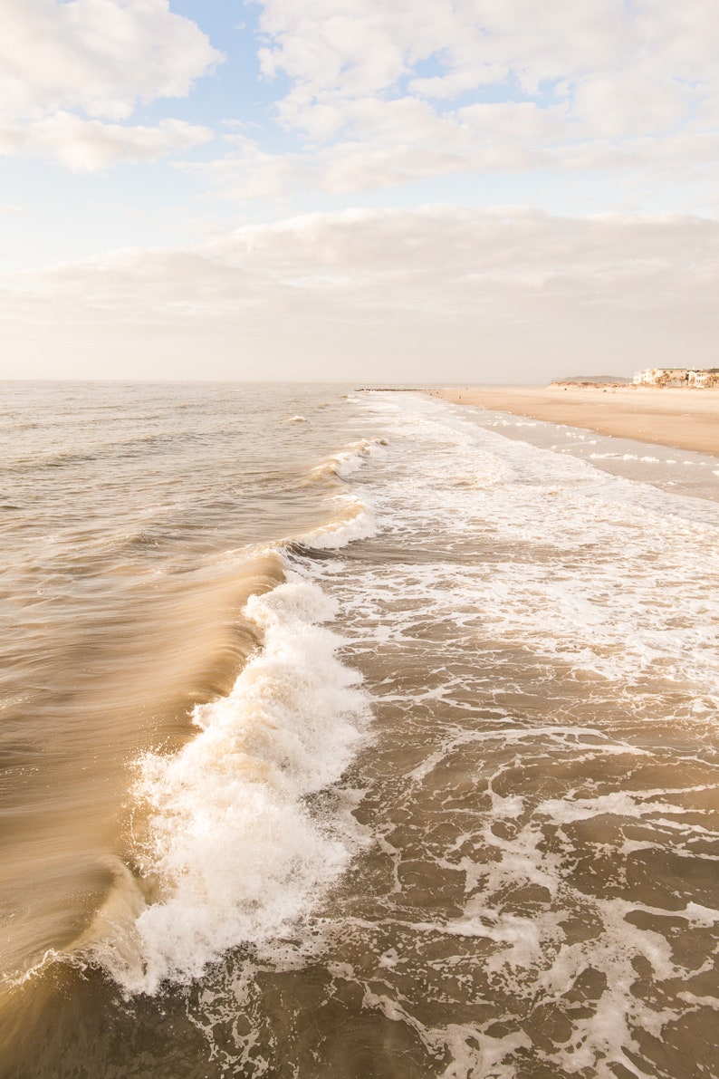 Pu&ograve; includere: Una serena scena di spiaggia con un'onda che si infrange in primo piano. L'acqua &egrave; di un marrone tenue, in contrasto con la schiuma bianca. Il cielo &egrave; azzurro pallido con nuvole soffici e la sabbia &egrave; beige chiaro.