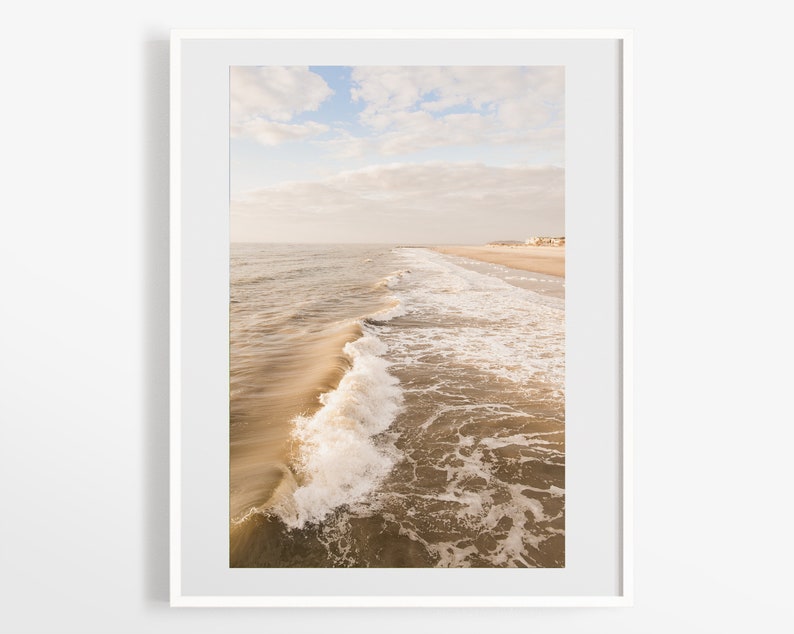 Pu&ograve; includere: Fotografia incorniciata di una scena di spiaggia. L'immagine cattura un'onda che si infrange vicino alla riva, con l'oceano e una spiaggia sabbiosa visibili. Il cielo &egrave; un mix di nuvole blu e bianche, creando un paesaggio costiero sereno.