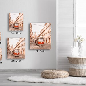 May include: A red and white tram travels down a narrow street lined with buildings in a European city. The street is cobblestone and the buildings are all beige with white trim. The tram is in focus and the buildings are slightly out of focus. The image is a photograph.