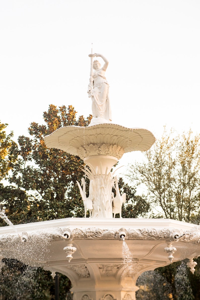 Pu&ograve; includere: Una fontana bianca e ornata con una statua di una donna che tiene una lancia in cima. L'acqua spruzza da pi&ugrave; bocche, creando un'esposizione rinfrescante. La fontana &egrave; circondata da una vegetazione lussureggiante, contro un cielo limpido.