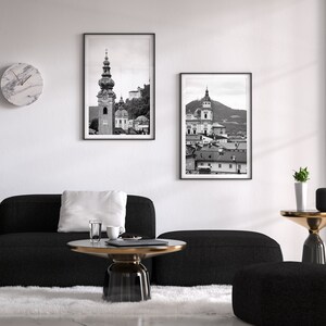 May include: Two framed monochrome prints of buildings hang above a black sofa and a round coffee table. A marble clock is on the wall. The room has a modern design with a white rug and a large window.