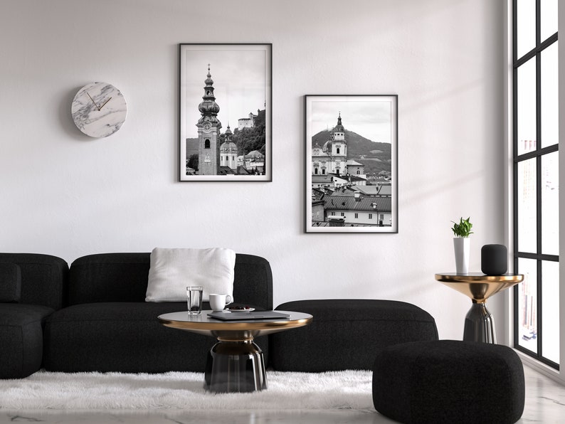 May include: Two framed monochrome prints of buildings hang above a black sofa and a round coffee table. A marble clock is on the wall. The room has a modern design with a white rug and a large window.