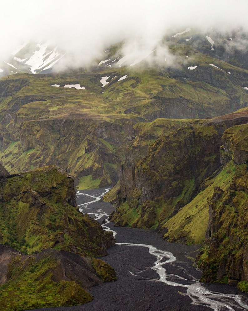 May include: A scenic landscape featuring a river winding through a deep canyon. The canyon walls are covered in green vegetation, with patches of snow on the distant mountains. Overcast skies and fog add to the dramatic atmosphere.
