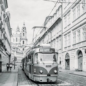 May include: A black and white photo of a vintage-style tram traveling down a cobblestone street in Prague, Czech Republic. The tram is numbered 8255 and has a sign that reads "22  Hradcany Stra&scaron;nice". The tram is traveling towards a church in the distance.