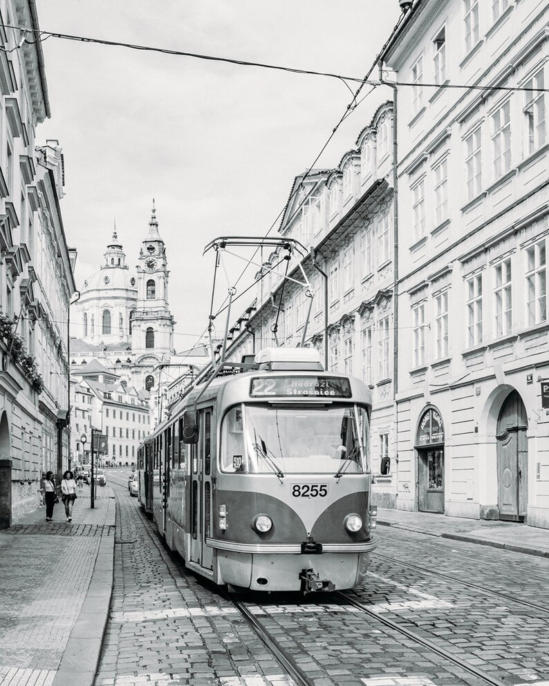 May include: A black and white photo of a vintage-style tram traveling down a cobblestone street in Prague, Czech Republic. The tram is numbered 8255 and has a sign that reads "22  Hradcany Stra&scaron;nice". The tram is traveling towards a church in the distance.