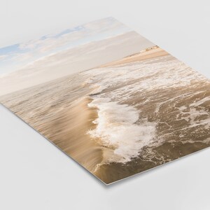 Pu&ograve; includere: Fotografia di una scena di spiaggia con vista prospettica. L'immagine cattura le onde dell'oceano mentre si muovono verso la riva. L'acqua &egrave; un mix di marrone e bianco, con il cielo azzurro pallido e beige.