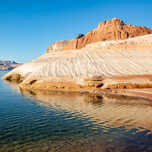 May include: A landscape photograph showing a lake with clear water reflecting the sky. The shoreline features layered sandstone formations in white, tan, and red hues. The sky is a clear, bright blue, typical of a sunny day.
