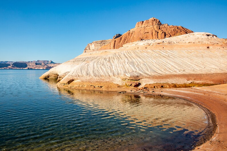 May include: A landscape photograph showing a lake with clear water reflecting the sky. The shoreline features layered sandstone formations in white, tan, and red hues. The sky is a clear, bright blue, typical of a sunny day.