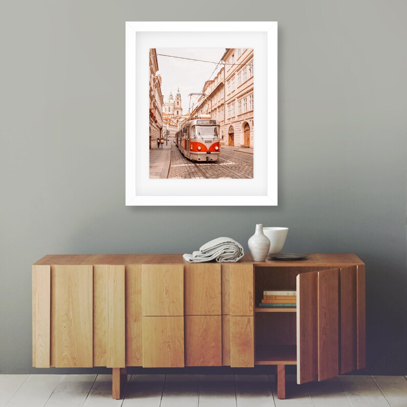 May include: A vintage-style red and silver tram travels down a cobblestone street in Prague, Czech Republic. The tram is surrounded by historic buildings with terracotta roofs.