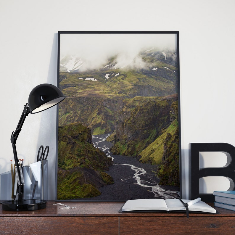 May include: A framed landscape photograph of a canyon with a river winding through it. The canyon walls are green and the riverbed is black. A black desk lamp, scissors, and a notebook are in the foreground.