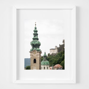 May include: Framed print of a European cityscape. The image shows a tall church spire with a green dome and cross, and a castle on a hillside. The sky is overcast, and the colour palette is muted.