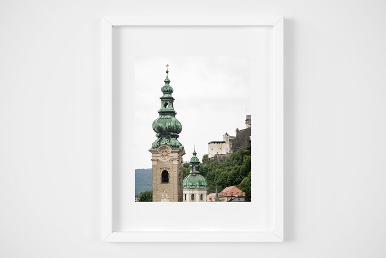 May include: Framed print of a European cityscape. The image shows a tall church spire with a green dome and cross, and a castle on a hillside. The sky is overcast, and the colour palette is muted.