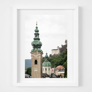 May include: A framed print depicting a European cityscape. The image shows a tall church tower with a green, onion-shaped dome and a clock. A castle is on a green hillside in the background, with other buildings visible.