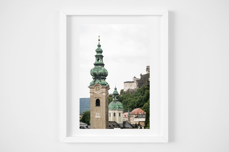 May include: A framed print depicting a European cityscape. The image shows a tall church tower with a green, onion-shaped dome and a clock. A castle is on a green hillside in the background, with other buildings visible.