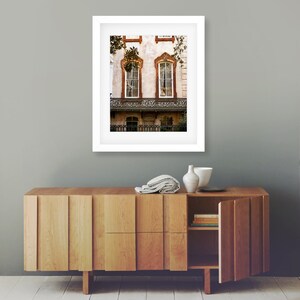 May include: A framed photograph of a historic building with two arched windows and a wrought iron balcony. The building has a light-colored facade with decorative trim. The photograph is displayed above a wooden cabinet with a partially open door.