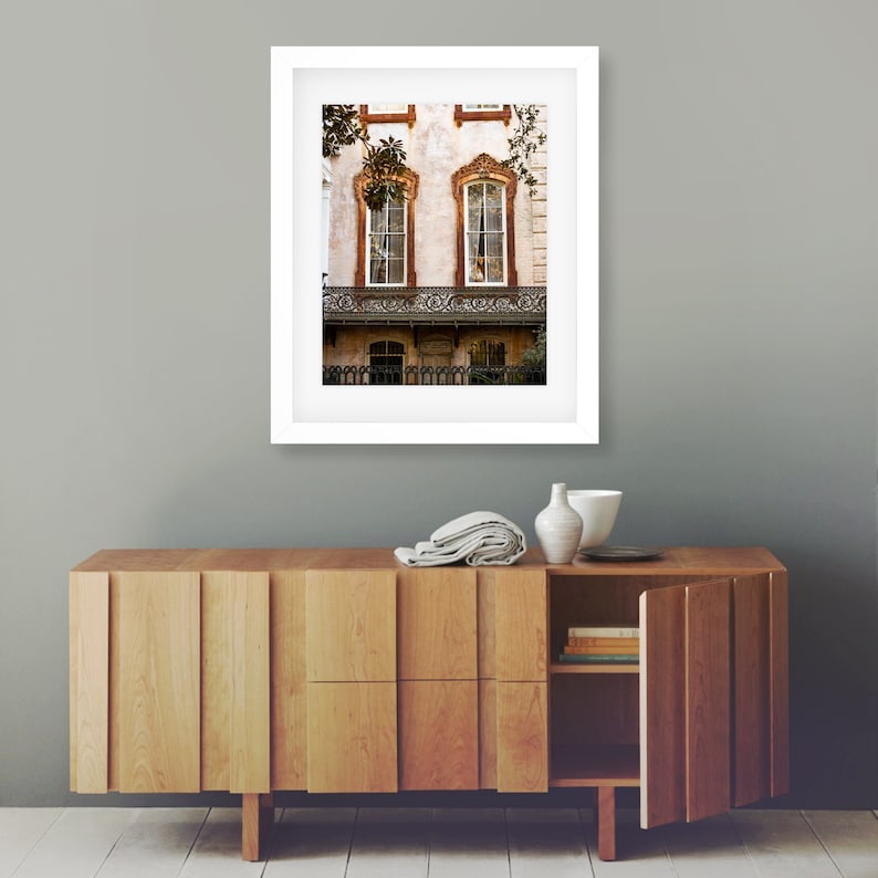 May include: A framed photograph of a historic building with two arched windows and a wrought iron balcony. The building has a light-colored facade with decorative trim. The photograph is displayed above a wooden cabinet with a partially open door.