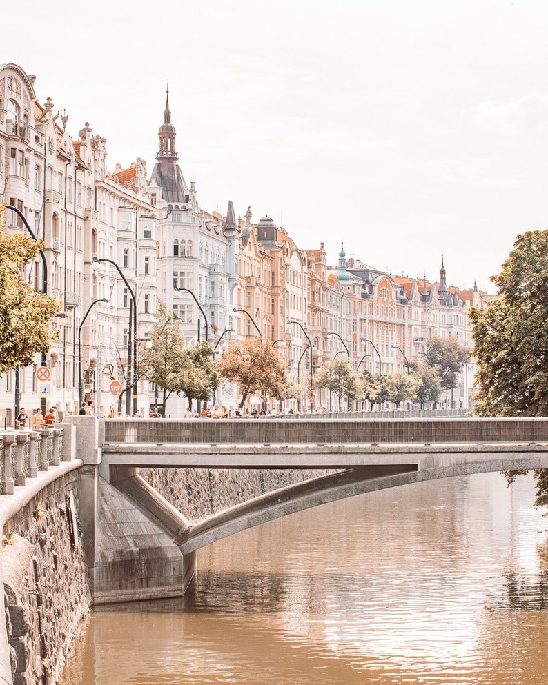 May include: A view of a bridge over a river in Prague, Czech Republic. The bridge is made of concrete and has a stone arch. The river is brown and the water is calm. The buildings on the other side of the river are tall and have colorful facades.