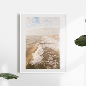 Pu&ograve; includere: Una stampa incorniciata con una serena scena di spiaggia. La fotografia cattura le onde dell'oceano che si infrangono sulla riva sotto un cielo nuvoloso. La tavolozza dei colori &egrave; morbida, con toni caldi di beige e blu, creando un'estetica rilassante.