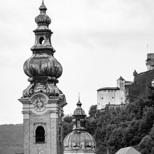 May include: Monochrome image of a European townscape, showcasing a church tower with a clock and intricate design. A domed building and a castle on a hill are also in view, set against a cloudy sky.