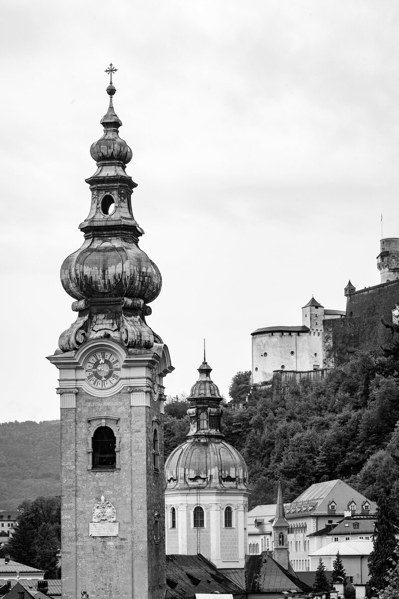 May include: Monochrome image of a European townscape, showcasing a church tower with a clock and intricate design. A domed building and a castle on a hill are also in view, set against a cloudy sky.