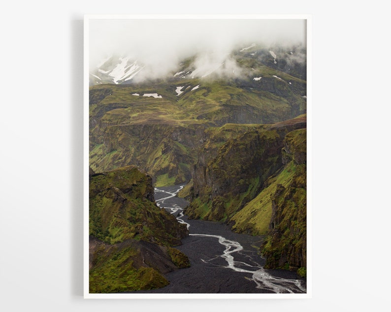 May include: A framed landscape print featuring a winding river through a deep canyon. The canyon walls are covered in green vegetation, with patches of snow on the distant mountain peaks. Overcast skies and fog add to the dramatic scenery.