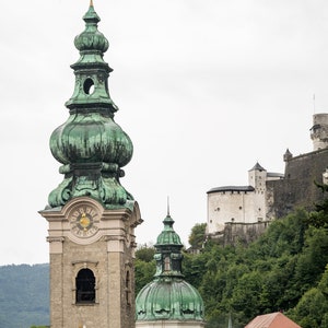 May include: A stone church tower with a green, onion-shaped dome and a clock face. The tower is part of a complex of buildings, including a castle on a hillside, and other buildings with red tile roofs. The sky is overcast.