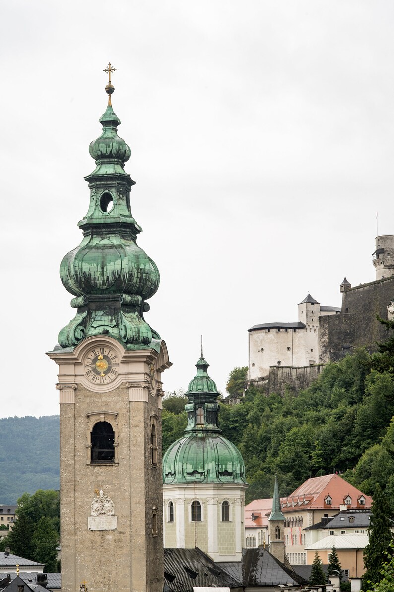 May include: A stone church tower with a green, onion-shaped dome and a clock face. The tower is part of a complex of buildings, including a castle on a hillside, and other buildings with red tile roofs. The sky is overcast.