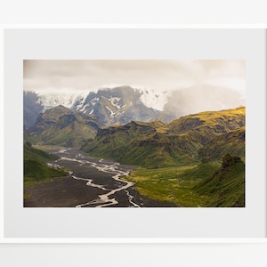 May include: A winding river flows through a valley surrounded by green and brown mountains. The mountains are covered in snow and clouds.