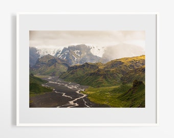 May include: A winding river flows through a valley surrounded by green and brown mountains. The mountains are covered in snow and clouds.