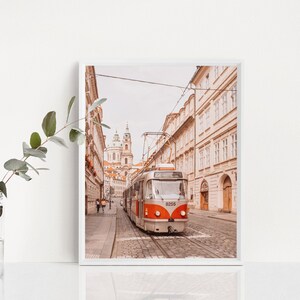 May include: A vintage-style tram travels down a cobblestone street in Prague, Czech Republic. The tram is orange and white with the number 8255 displayed on the front. The street is lined with tall buildings in a warm, muted color palette.