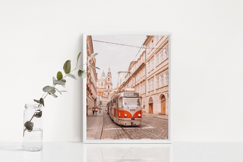 May include: A vintage-style tram travels down a cobblestone street in Prague, Czech Republic. The tram is orange and white with the number 8255 displayed on the front. The street is lined with tall buildings in a warm, muted color palette.