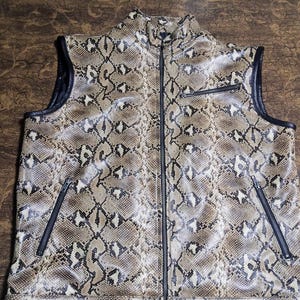 May include: A sleeveless vest featuring a brown, black, and cream snakeskin pattern. The garment has a high collar, a full-length central zipper, two slanted zippered pockets at the waist, and one zippered chest pocket, all with black trim.