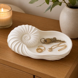 Könnte beinhalten: Eine weiße Schmuckschale mit matter Oberfläche in Form eines Nautilus-Gehäuses. Die Schale hat ein spiralförmiges Muscheldesign auf einer Seite und eine glatte, flache Vertiefung für kleine Gegenstände. Darin liegen zwei Goldringe, eine Kette und ein Schlüssel.