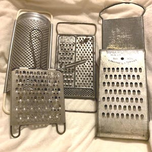 May include: A collection of four vintage metal kitchen graters. One features a white frame, while another is labeled "ALL-IN-ONE PAT. PEND." and a third is marked "WONDER SHREDDER REG. U.S. PAT. OFF. PAT. NOS. 1.768.482 & 1.768.483" with perforated surfaces.